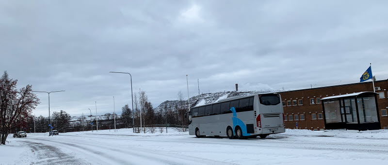 Fotot visar en grå buss på en snöklädd gata vid en busshållplats.