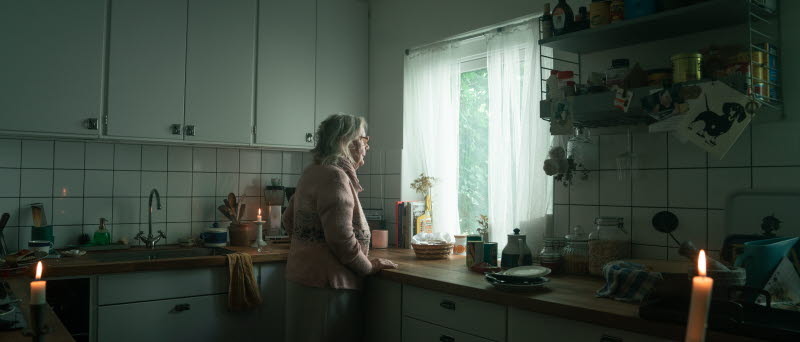 Äldre kvinna står i ett mörkt kök med tända stearinljus. Hon tittar ut genom fönstret.
