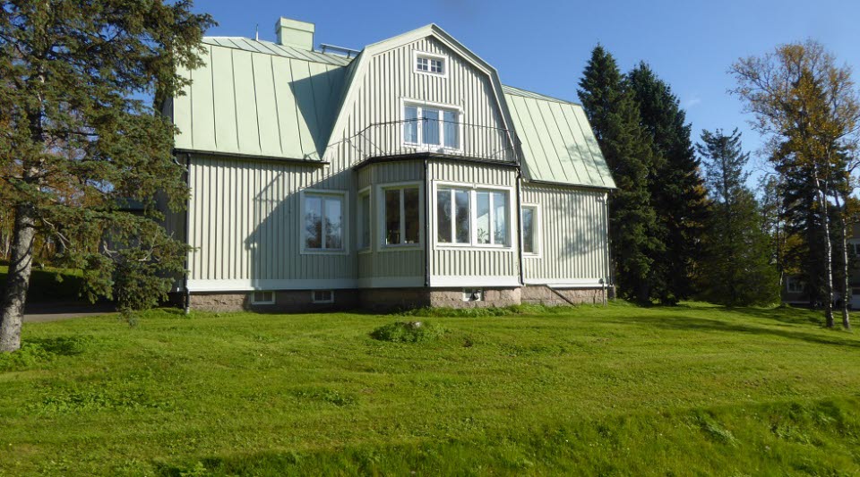 Fotot visar ett grått stort trähus med grönt tak. Bakom huset ses gröna träd och framför en grön gräsmatta.