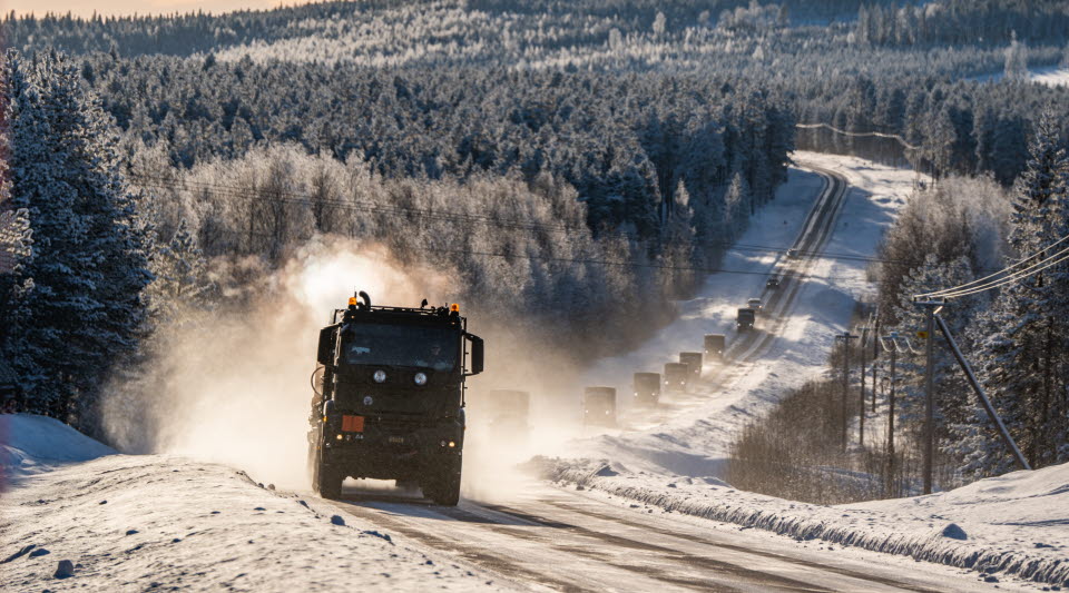 En militärkolonn kör längs en väg med snörök i vintrig miljö.