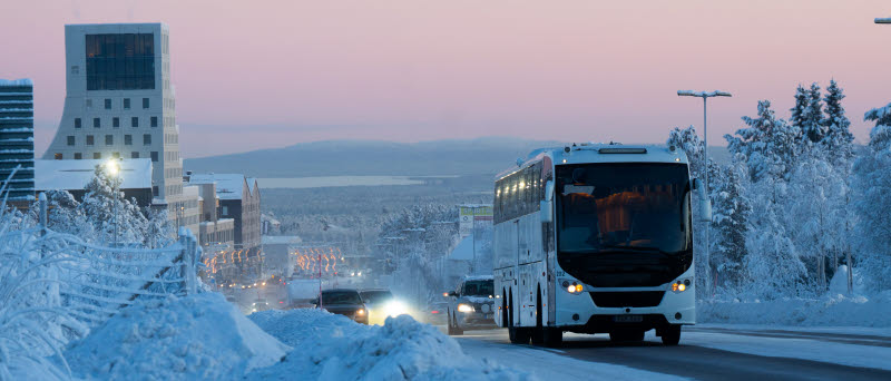 Buss kör på en vinterväg genom en snötäckt stad i skymning.