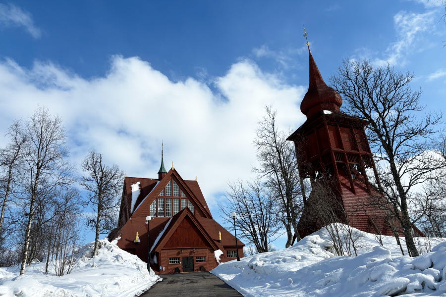 Fotot visar kyrkan med klockstapel omgiven av snöklädd mark mot en blå himmel med delvis moln. 