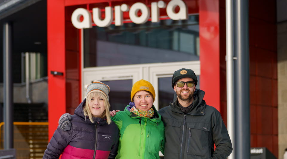 Tre personer i färgglada kläder som håller varandra om axlarna utanför kulturhuset Aurora i Kiruna