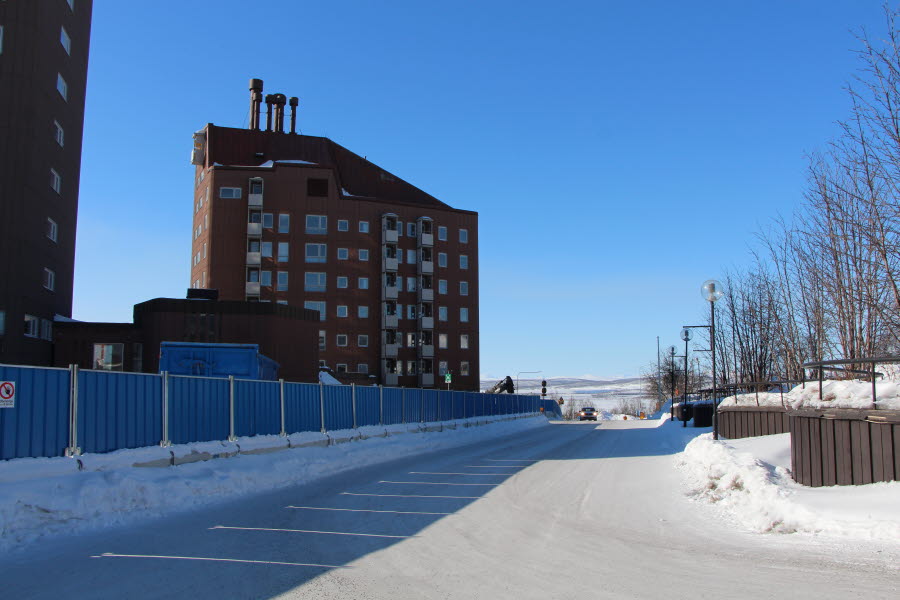 Fotot visar ett brunt höghus bakom ett stängsel och en väg bredvid.