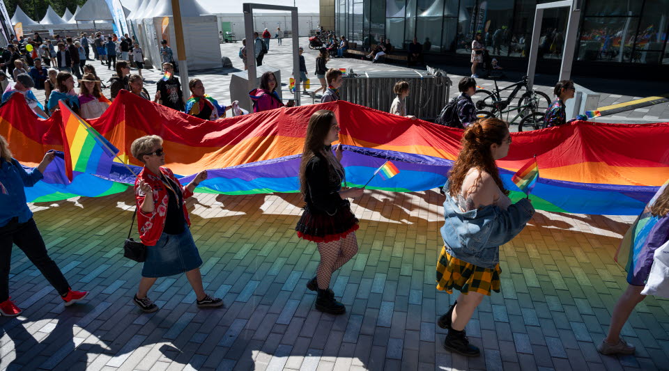 Flera människor paraderar med prideflaggan. De håller i den tillsammans. Det är sommar. I bakgrunden syns Stadshustorget i Kiruna.