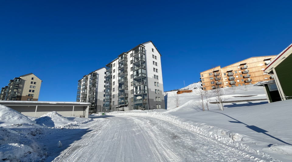Fotot visar en blandning av höga bostadshus, lägre bostadshus och träbyggnader i ett bostadsområde vintertid med blå himmel som bakgrund. 