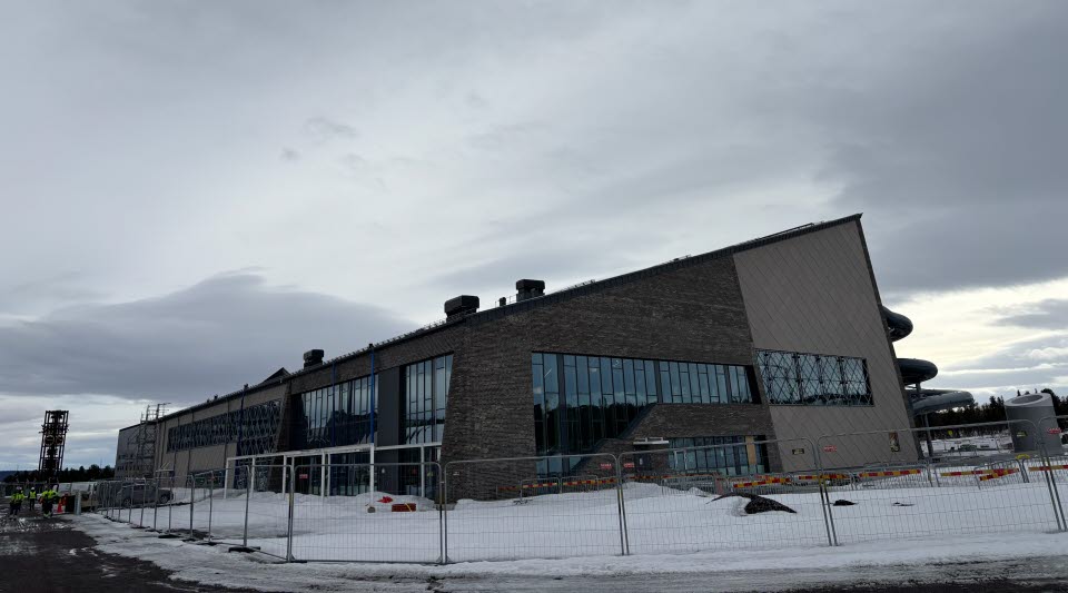 Fotot visar en grå byggnad med stängsel runt och del av en stadskärna bakom. 