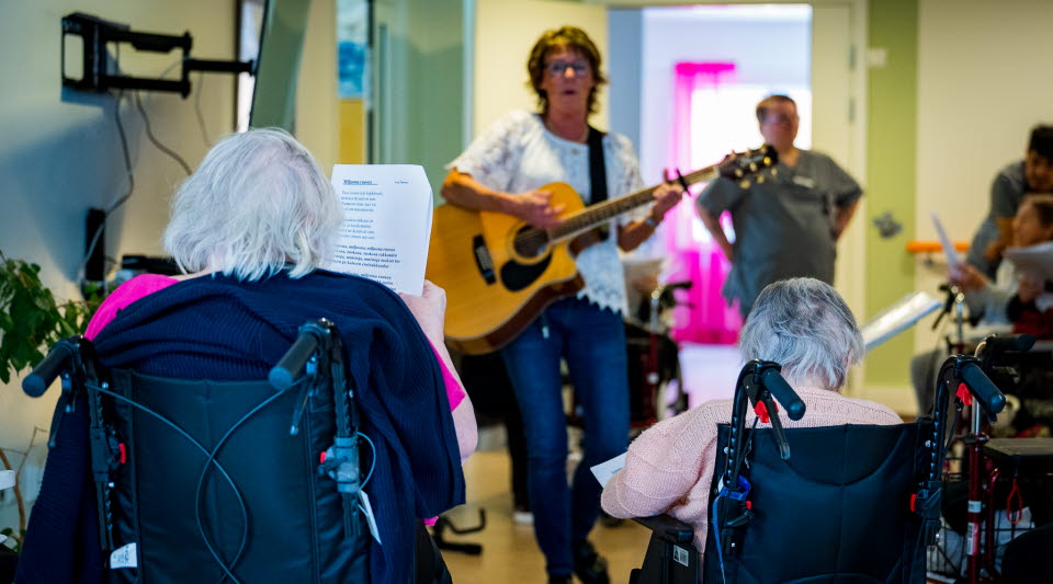 Äldre personer, sittandes i rullstol sjunger tillsammans med en allsångsledare med gitarr. 