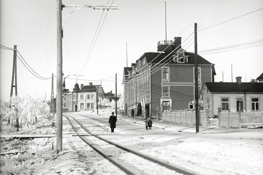 Gammal vintergata i Kiruna med spårväg, elstolpar och människor som promenerar mellan träbyggnader, historisk stadsmiljö, svartvitt foto.