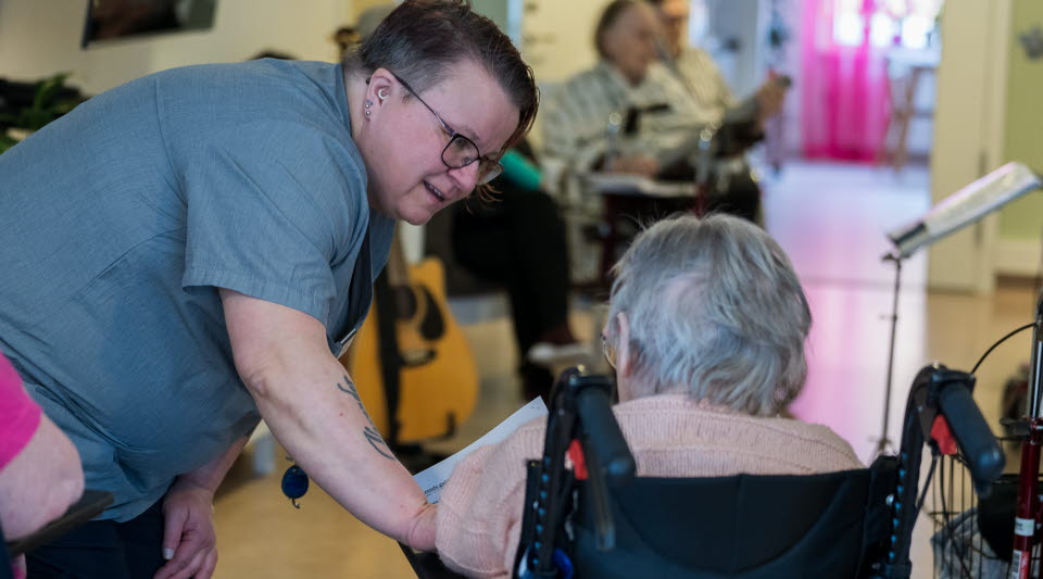 Ett vårdbiträde som leende vänder sig mot en äldre person sittandes i rullstol.