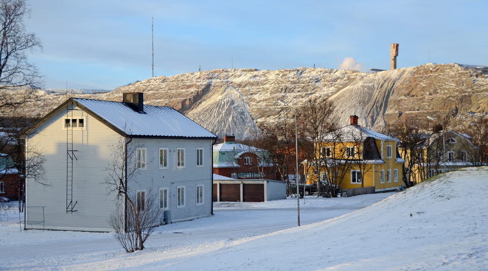 Fotot visar ett bostadsområde med gamla trähus i olika färger på vintern med ett snöklätt berg bakom. 