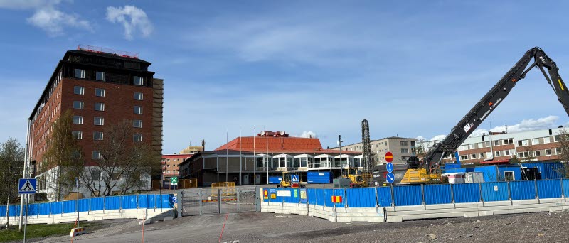 Gamla Scandic Ferrum och Folkets hus inramade av blå staket. En traktor står och gräver till vänster om byggnaderna. I början av juni.
