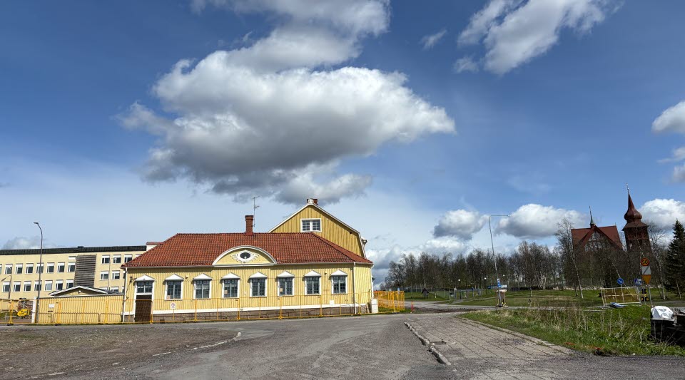 Fotot visar en gul byggnad bakom stängsel och en kyrka med klocktorn till höger. 