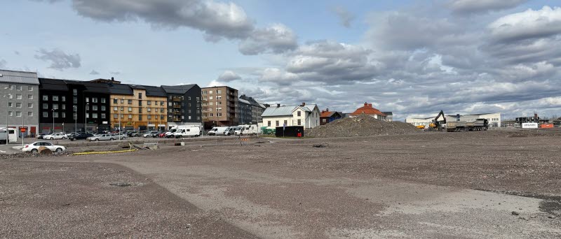 Fotot visar en öppen plats där markarbete pågår med en huslänga i bakgrunden.