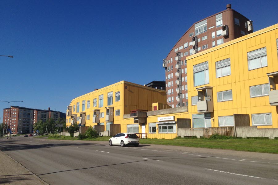 Fotot visar en väg med gula låga byggnader och en brun högre byggnad bakom mot en blå himmel. 