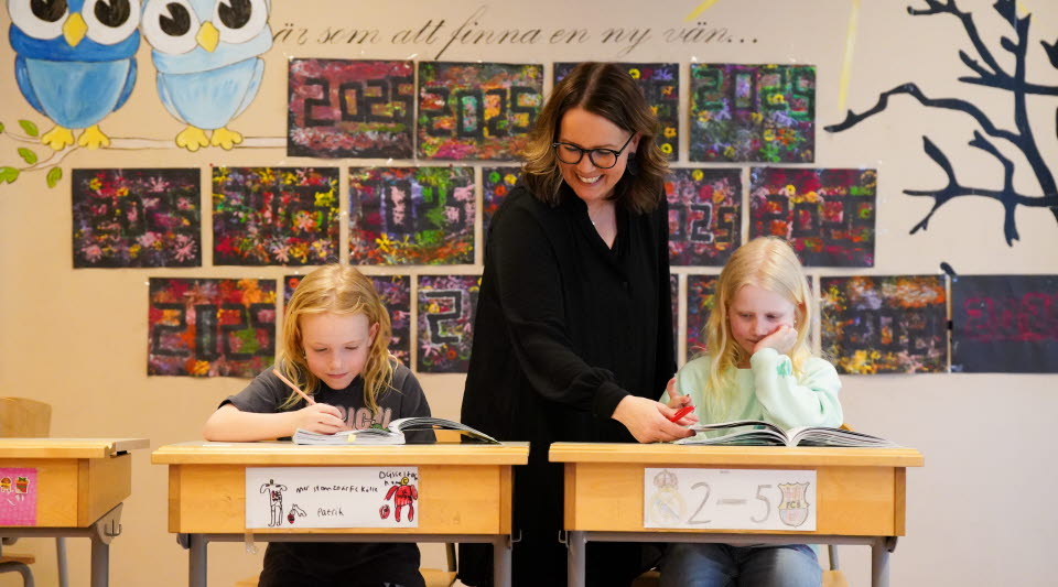 Två barn sitter i skolbänkar och skriver, leende lärare står bredvid. 