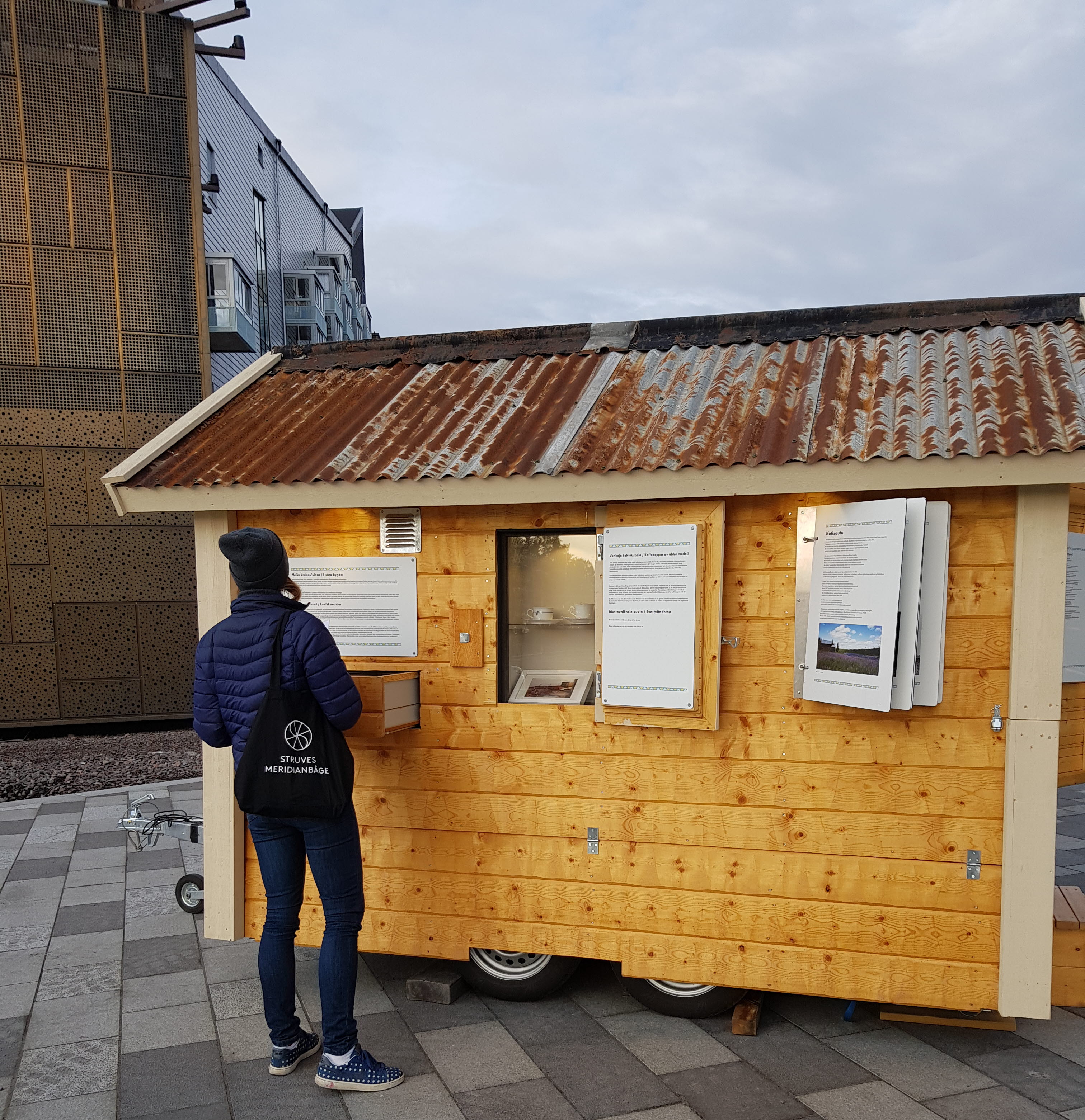Fotot visar en träfärgad byggnad med informationsblad och en person som står och läser 