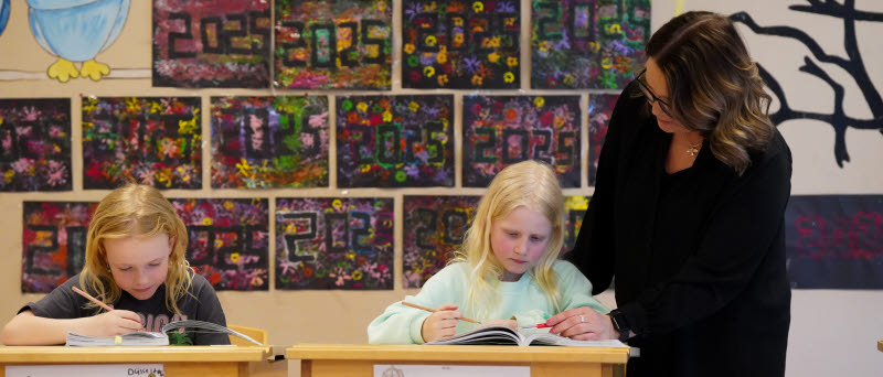 Två barn sitter och skriver vid sina bänkar. Ett av barnen får hjälp av en lärare.