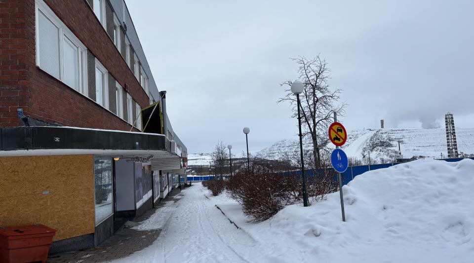 Fotot visar en snöklädd trottoar med ett blått stängsel och en gruva i bakgrunden. 