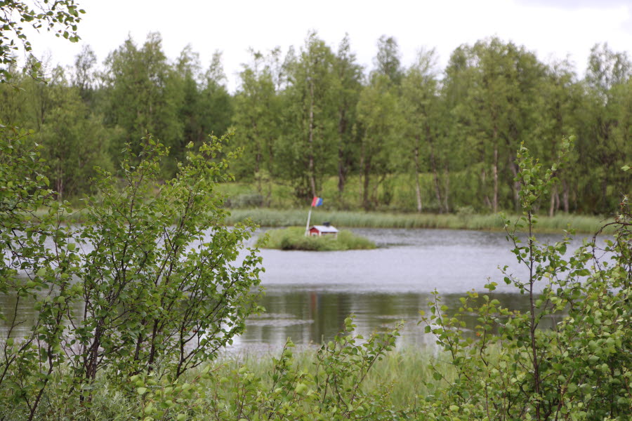 Fotot är taget på sommaren och visar en liten röd stuga på en ö med gröna träd runt omkring. 
