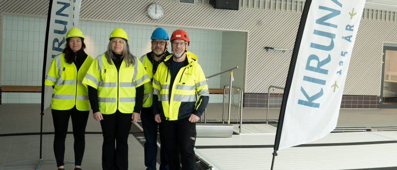 Fotot visar fyra personer klädda i varselkläder vid beachflaggor i ett badhus.