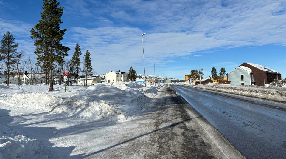 Fotot visar en väg med hus på sidorna och snöklädd mark med blå himmel i bakgrunden.