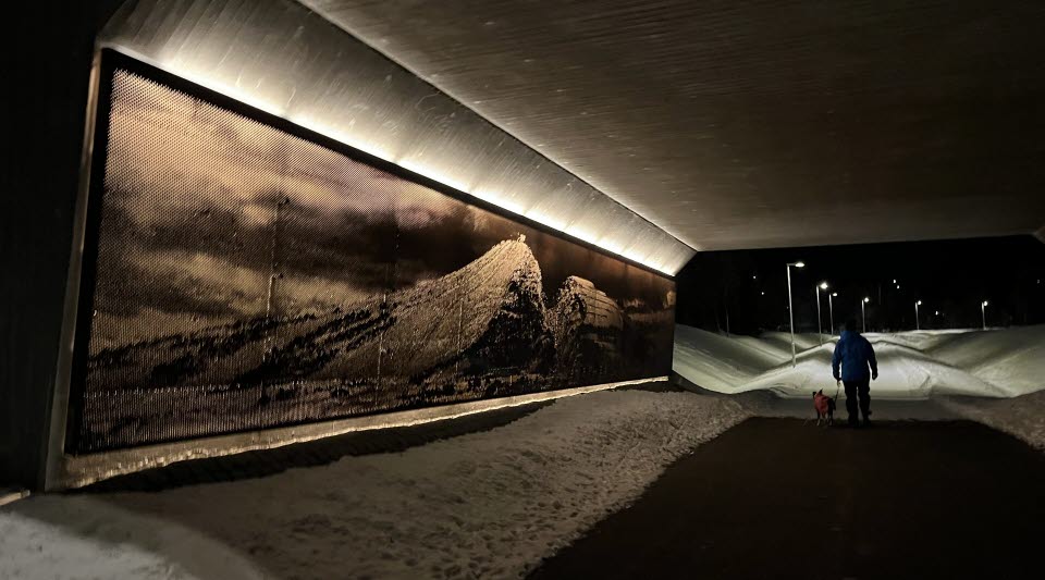 Fotot är taget på kvällen och visar en viadukt med en ljusinstallation av fjället Luossavaara på ena sidan och en person med hund på gångvägen. .