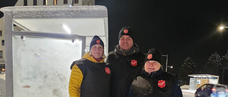 Två kvinnor och en man står framför en busskur i vintermiljö.