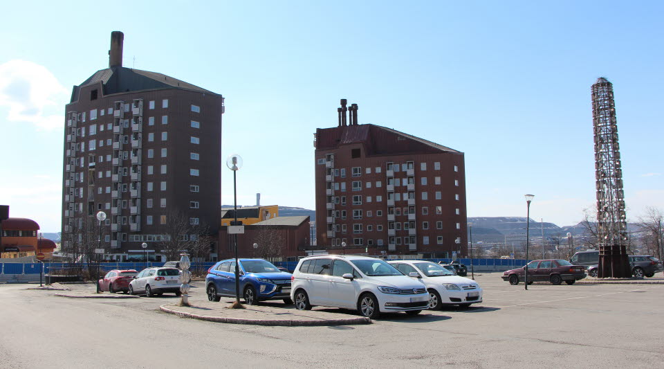 Fotot visar en parkeringsplats med bilar och hus i bakgrunden.