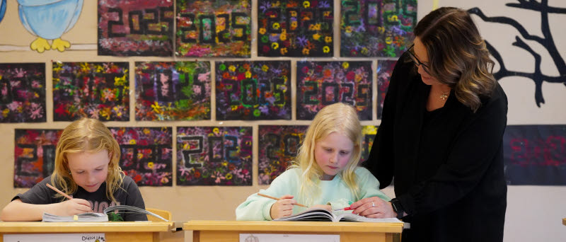 Två elever som sitter vid sina skolbänkar och skriver i sina böcker. En lärare står bredvid och hjälper till.
