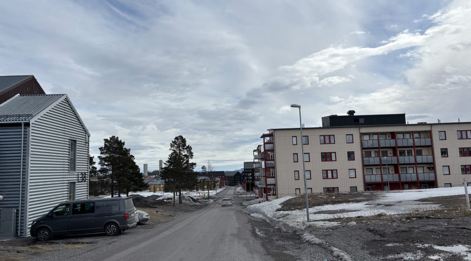 Fotot visar en gata med hus i olika färger, parkerade bilar, och fläckar med snö på marken.
