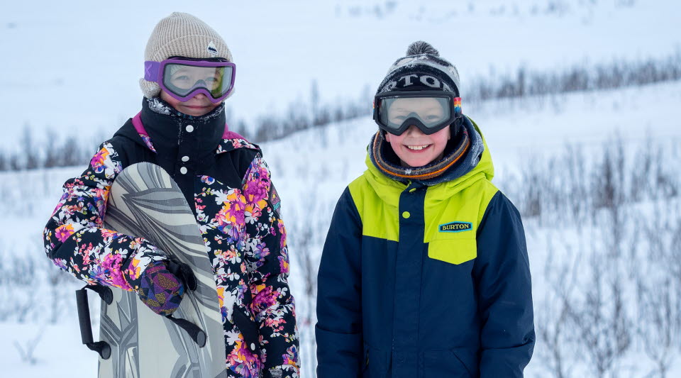 Två barn i en pulkabacke, klädda i vinterkläder och slalomglasögon. Ena håller i en åkmadrass. 