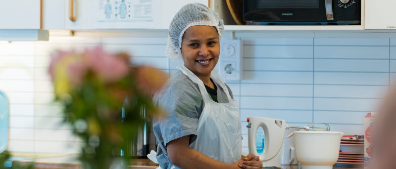 Fotot visar en person som jobbar i ett kök iklädd vita och blå arbetskläder.