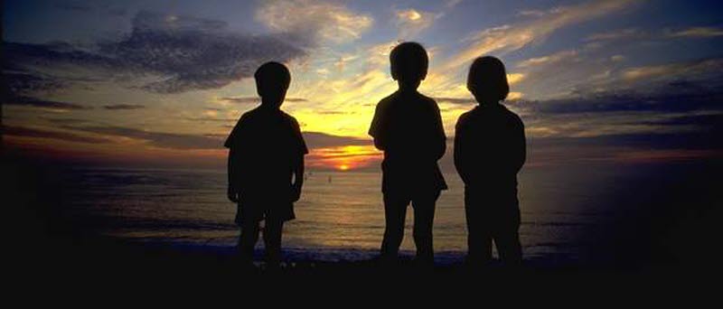 Tre barn står i skymningen med ryggarna mot kameran och blickar ut över en solnedgång.
