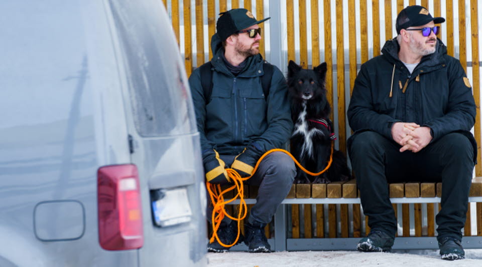 Två personer och en hund sitter och väntar på bussen i en busskur